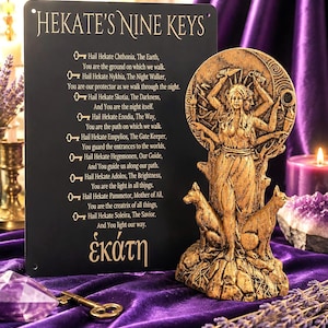 May include: A gold-colored statue of Hecate with three faces and multiple arms, flanked by two dogs, stands on a textured base. A black sign with the text "HEKATE'S NINE KEYS" and a list of invocations is to the left. The word "ἑκάτη" is at the bottom.