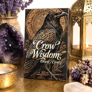 Op de afbeelding: Een rechthoekig kaartspel Crow Wisdom Oracle Cards met een zwarte kraai-illustratie tegen een gouden mandala-achtergrond. De tekst Crow Wisdom Oracle Cards staat in witte schrijf- en schreefletters op de voorkant van de zwarte doos.