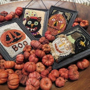 May include: Four framed Halloween wall hangings in black frames, depicting a black cat, jack-o'-lanterns, and other spooky characters. The hangings are surrounded by small, orange pumpkins in a wooden bowl, creating an autumnal display.