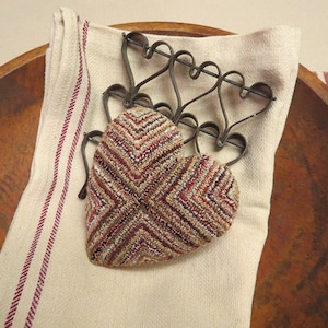 May include: A brown and white heart-shaped knitted pillow with a red and white striped pattern sits on a white linen towel with red stripes. The pillow is resting on a wooden bowl.