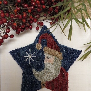 Op de afbeelding: Een handgestikte kerstversiering in de vorm van een ster met een afbeelding van de kerstman. Het ontwerp maakt gebruik van blauw, rood, beige en wit garen om het gezicht van de kerstman en een sneeuwvlok te creëren. De achtergrond is een crèmekleurige stof.