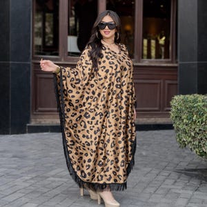 May include: A long, flowing caftan in a tan and black leopard print. The caftan has a V-neck and is trimmed with black fringe along the hem and sleeves. The person is wearing sunglasses and heels.
