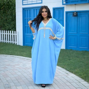 Op de afbeelding: Een lichtblauwe kaftan jurk met gouden afwerking rond de halslijn en mouwen. De jurk heeft een los, vloeiend ontwerp. De vrouw op de foto draagt zwarte sandalen en gouden sieraden.