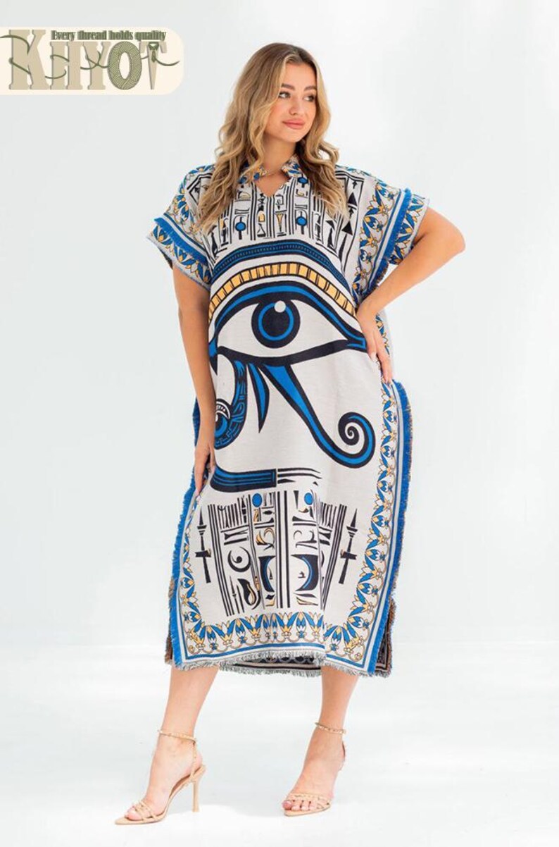May include: A white, knee-length caftan dress with a blue and gold Eye of Horus design. The dress has short sleeves and a split hem with blue fringe. The text "Every thread holds quality" is at the top left.