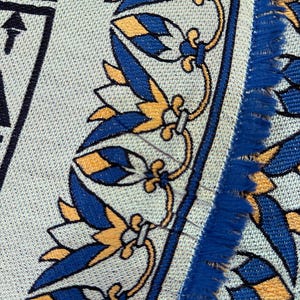 May include: Close-up of a textile with a repeating pattern of blue and gold floral and geometric designs on a light gray background. The edge has a blue fringe. The text "Every thread holds quality" is visible.