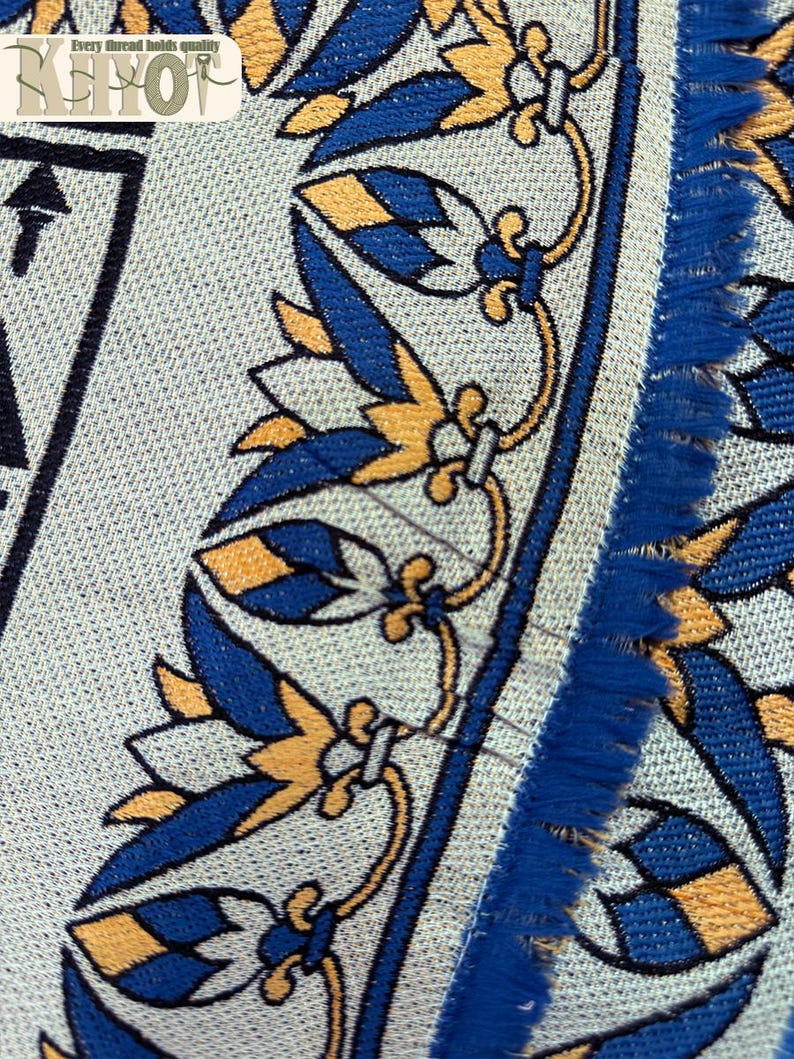 May include: Close-up of a textile with a repeating pattern of blue and gold floral and geometric designs on a light gray background. The edge has a blue fringe. The text "Every thread holds quality" is visible.