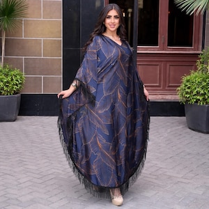 May include: A navy blue caftan dress with a V-neck and flowing sleeves. The dress features a repeating pattern of gold-colored leaf designs. Black fringe trims the sleeves and hemline. The dress is worn with beige shoes.