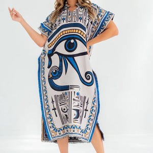 May include: A knee-length, white caftan dress with a blue and gold Egyptian eye of Horus design. The dress has short sleeves, a fringed hem, and a split on the side. The text "Every thread holds quality" is at the top.