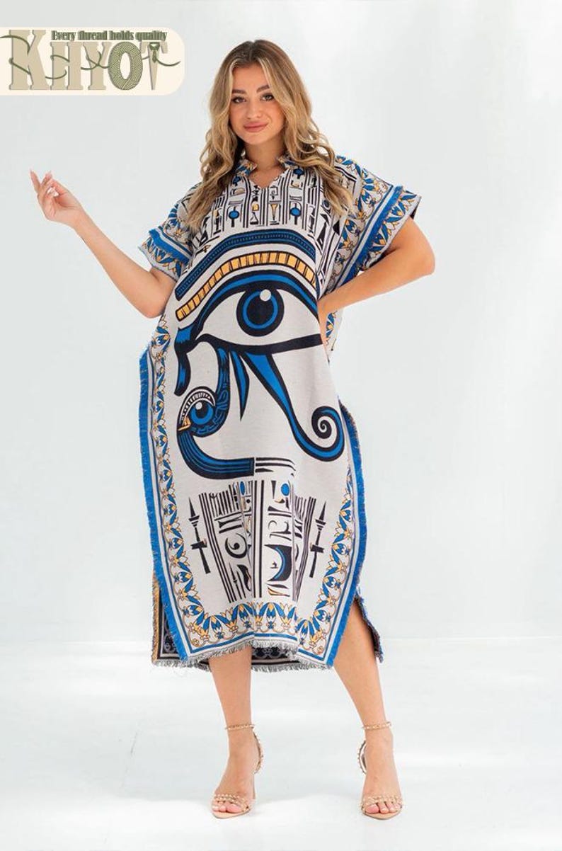 May include: A knee-length, white caftan dress with a blue and gold Egyptian eye of Horus design. The dress has short sleeves, a fringed hem, and a split on the side. The text "Every thread holds quality" is at the top.