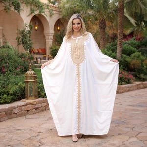 May include: A white caftan with gold embroidery around the neckline and down the front. The long-sleeved garment is made of a flowing fabric. The caftan is worn outdoors in a garden setting.