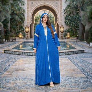 May include: A long, royal blue hooded dress with white and gold trim. The dress features long sleeves and a vertical white stripe down the front. The woman is standing in front of a fountain and archway.