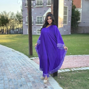 May include: A vibrant purple pleated outfit, featuring a long, flowing top with fringe trim and matching pants. The outfit has a V-neck design and is paired with beige platform heels. The ensemble is suitable for special occasions.