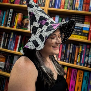 May include: A tall, pointed hat featuring a quilted patchwork design in black, white, and purple. The exterior displays a geometric pattern, while the underside of the wide, floppy brim is black with a white spiderweb print throughout the fabric.