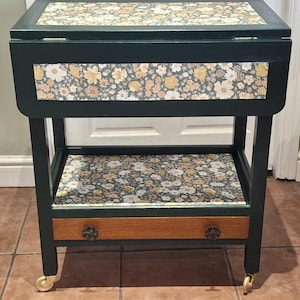 May include: A dark green tea trolley with a floral patterned top, shelf, and front panel. The trolley has a wooden drawer with decorative knobs and gold-coloured caster wheels. The floral pattern includes white, yellow, and peach-coloured flowers.