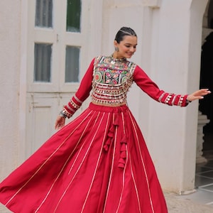 Può includere: Un vivace completo rosso con un top a maniche lunghe con ricami intricati e una gonna lunga abbinata. La gonna ha strisce bianche verticali. Il top presenta dettagli elaborati e la gonna ha un dettaglio con nappe.