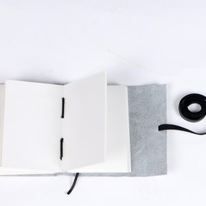 May include: A gray leather-bound journal with blank pages is open and lying flat on a white surface. A black ribbon is tied around the journal and a black ribbon is coiled on the surface.
