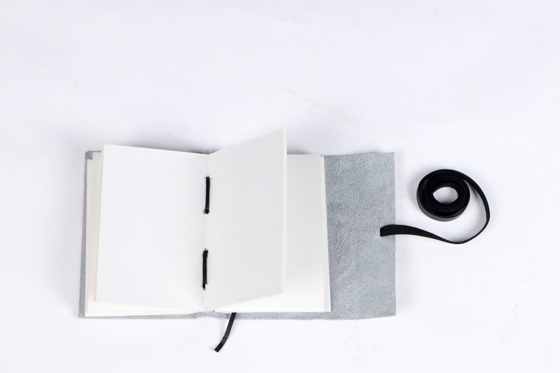 May include: A gray leather-bound journal with blank pages is open and lying flat on a white surface. A black ribbon is tied around the journal and a black ribbon is coiled on the surface.