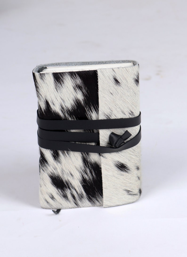 May include: A black and white cowhide journal with a black leather strap and a bow tie closure.