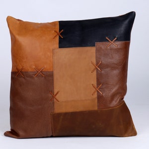 May include: Square leather cushion with a patchwork design. The cushion features various shades of brown and a black square, with decorative cross-stitch details. A decorative home accent.