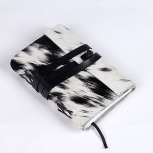 May include: A black and white cowhide journal with a black leather strap closure. The journal has a textured surface and a simple design.