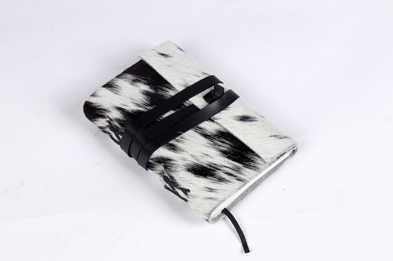 May include: A black and white cowhide journal with a black leather strap closure. The journal has a textured surface and a simple design.