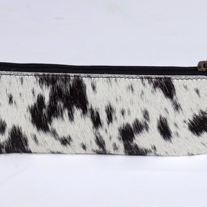 Puede incluir: Una bolsa de cremallera de piel de vaca en blanco y negro. La bolsa está hecha de piel suave y flexible con una cremallera negra y una tira de cuero negra.