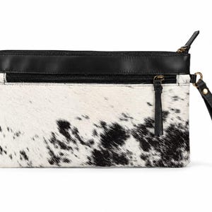 Op de afbeelding: Een rechthoekige clutch met een zwart-wit koeienhuidpatroon op het hoofdgedeelte. Het bovenste gedeelte is gemaakt van glad zwart leer met een horizontale ritszak. Het bevat een zwarte leren polsband die aan de zijkant is bevestigd.