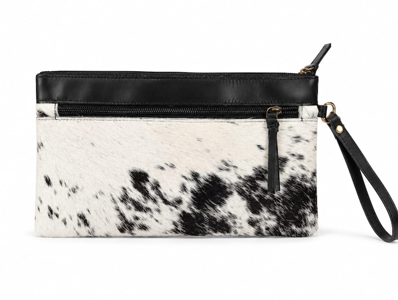 Op de afbeelding: Een rechthoekige clutch met een zwart-wit koeienhuidpatroon op het hoofdgedeelte. Het bovenste gedeelte is gemaakt van glad zwart leer met een horizontale ritszak. Het bevat een zwarte leren polsband die aan de zijkant is bevestigd.