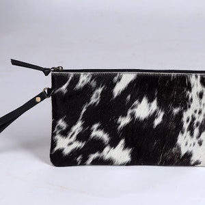 Op de afbeelding: Een rechthoekige clutch met een zwart-wit koeienhuidpatroon en een textuur van echt haar. De tas is voorzien van een zwarte ritssluiting en een bijpassende zwarte lederen polsband die aan de zijkant is bevestigd met een goudkleurige metalen klinknagel.