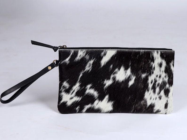 Op de afbeelding: Een rechthoekige clutch met een zwart-wit koeienhuidpatroon en een textuur van echt haar. De tas is voorzien van een zwarte ritssluiting en een bijpassende zwarte lederen polsband die aan de zijkant is bevestigd met een goudkleurige metalen klinknagel.