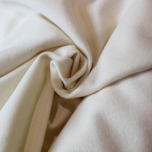 May include: A piece of off-white fabric with a soft, matte texture and a dense, felt-like appearance. The material is gathered into a central swirl, creating deep, rounded folds and shadows that highlight the smooth, uniform surface of the textile.