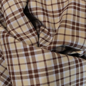 May include: Close-up of a fabric with a brown and cream plaid pattern. The fabric is folded and draped, showing the texture and design. The plaid pattern consists of intersecting lines forming squares.