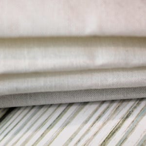 May include: A stack of four white fabric squares, folded neatly, with a light gray fabric square underneath. The fabric squares are on a striped surface.