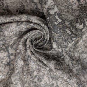 May include: A close-up of a fabric with a swirling pattern. The fabric is a blend of gray and beige, with darker gray, tree-like designs. The fabric is soft and appears to be draped, with a spiral in the center.