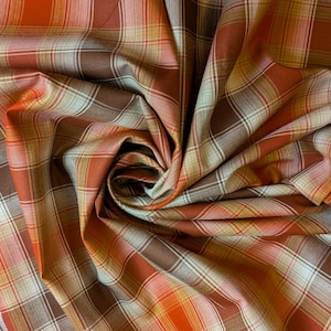 May include: Close-up of a plaid fabric with a mix of warm colors. The pattern features a grid of intersecting lines in shades of orange, brown, tan, and white. The fabric is folded and draped, creating shadows and depth.