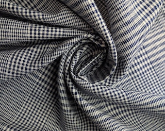 May include: Close-up of a fabric with a classic glen plaid pattern. The fabric features a navy blue and white check design, with the fabric folded and draped to show texture and dimension. Ideal for apparel and home decor.