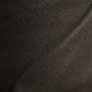 May include: Close-up of a dark gray fabric with a textured surface. The fabric appears to be woven, with subtle variations in tone and texture. The material is likely a textile, possibly wool or a similar material.