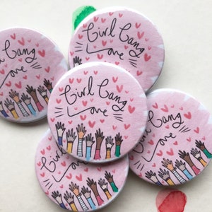 Può includere: Una collezione di bottoni rotondi rosa con la scritta "Girl Gang Love" in corsivo nero. I bottoni presentano mani alzate in varie tonalità di pelle e piccoli cuori rosa. Un accessorio carino e positivo.