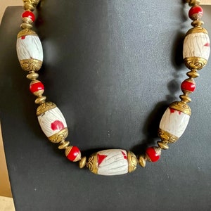 Puede incluir: Un collar de cuentas blancas y rojas con detalles dorados. Las cuentas son de forma ovalada y tienen una superficie texturizada. El collar está en un maniquí negro.