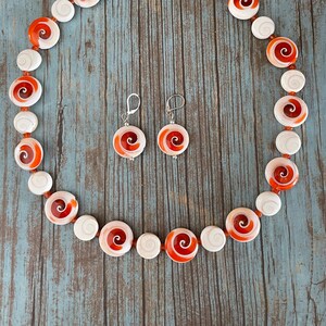 Peut inclure: Un ensemble de bijoux en coquillages rouge et blanc, comprenant un collier et des boucles d'oreilles. Le collier est composé de coquillages ronds et plats avec un motif en spirale. Les boucles d'oreilles sont également rondes et plates, avec un motif en spirale.