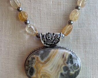 May include: A silver necklace with a large oval pendant featuring a brown and white agate stone. The necklace is made with small, round, light brown beads.