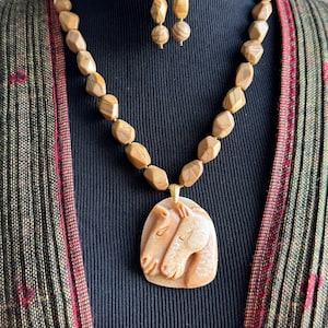 Puede incluir: Un conjunto de collar y pendientes con un colgante con dos cabezas de caballo talladas en piedra marrón claro. El collar tiene cuentas geométricas grandes de un color similar. Los pendientes tienen cuentas a juego y ganchos dorados.