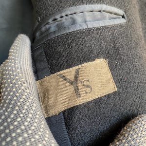 May include: Close-up of a dark gray wool coat interior. The coat features a small pocket with a light blue lining and a tan label with the letters "Y's" printed on it. The coat's interior is visible.