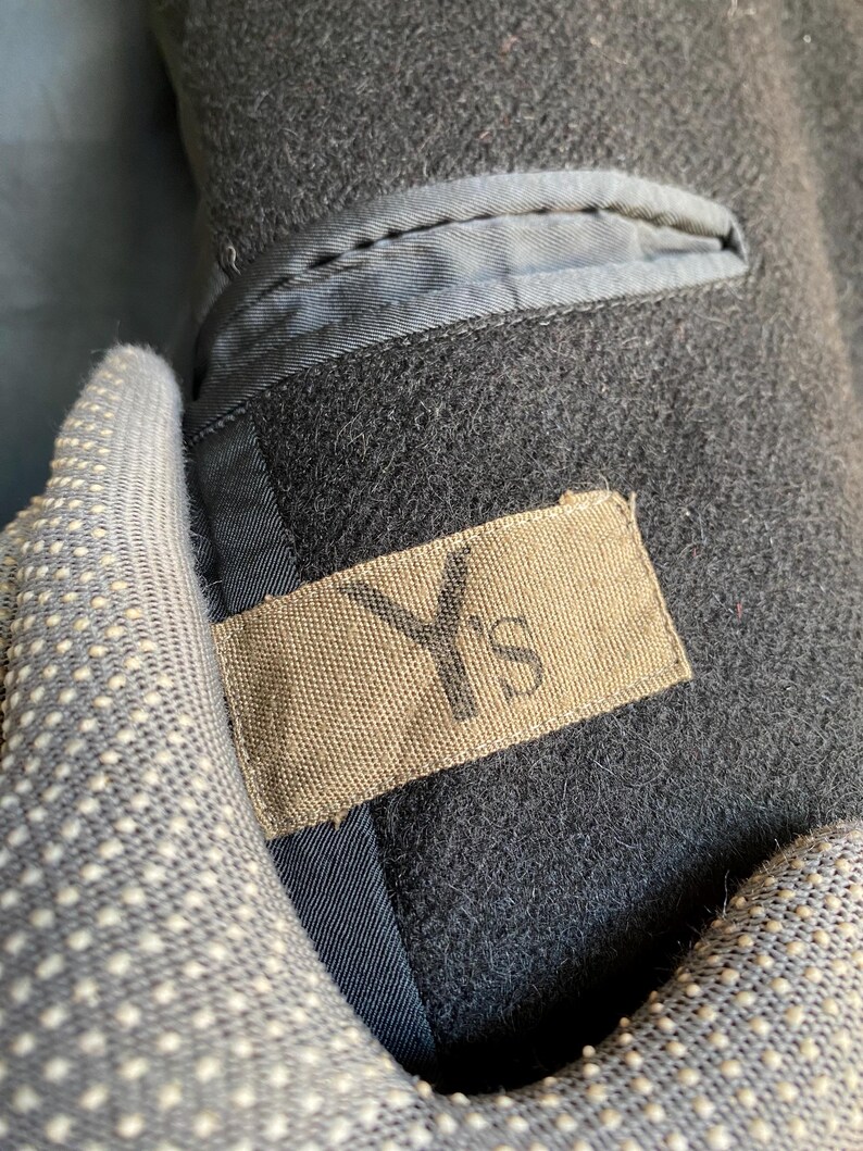 May include: Close-up of a dark gray wool coat interior. The coat features a small pocket with a light blue lining and a tan label with the letters "Y's" printed on it. The coat's interior is visible.