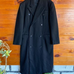 May include: A long, black double-breasted coat with six buttons and a notched lapel. The coat is hanging on a black hanger against a wooden background. The coat appears to be made of a heavy fabric.