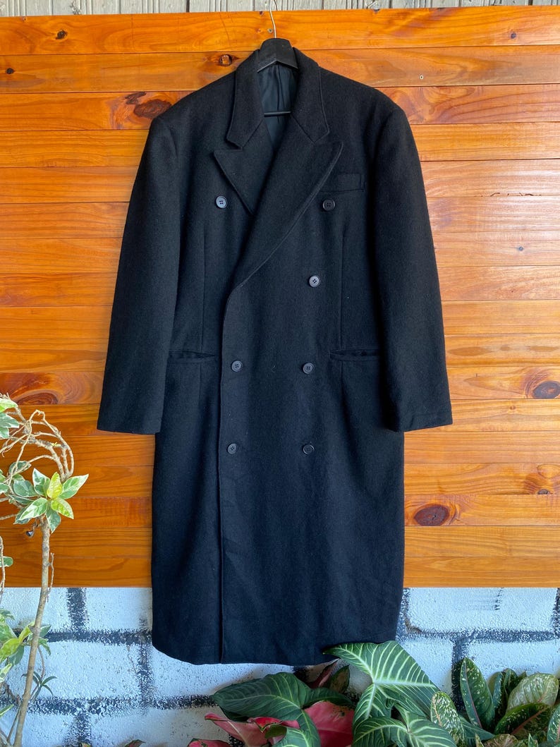 May include: A long, black double-breasted coat with six buttons and a notched lapel. The coat is hanging on a black hanger against a wooden background. The coat appears to be made of a heavy fabric.