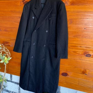 May include: A long, black double-breasted coat with six buttons and a notched lapel. The coat is hanging on a black hanger against a wooden background. The coat appears to be made of a heavy wool or wool blend.