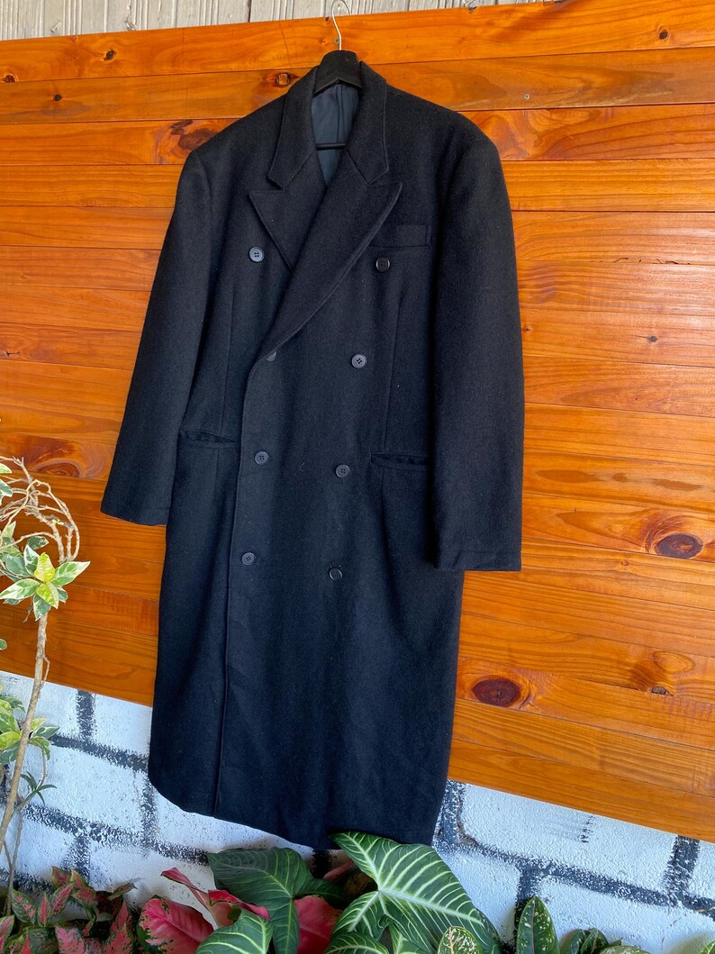 May include: A long, black double-breasted coat with six buttons and a notched lapel. The coat is hanging on a black hanger against a wooden background. The coat appears to be made of a heavy wool or wool blend.