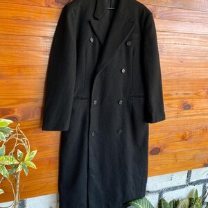May include: A long, black double-breasted coat with a classic design. The coat features a notched lapel, multiple buttons, and a tailored fit. It is hanging on a black hanger against a wooden background.