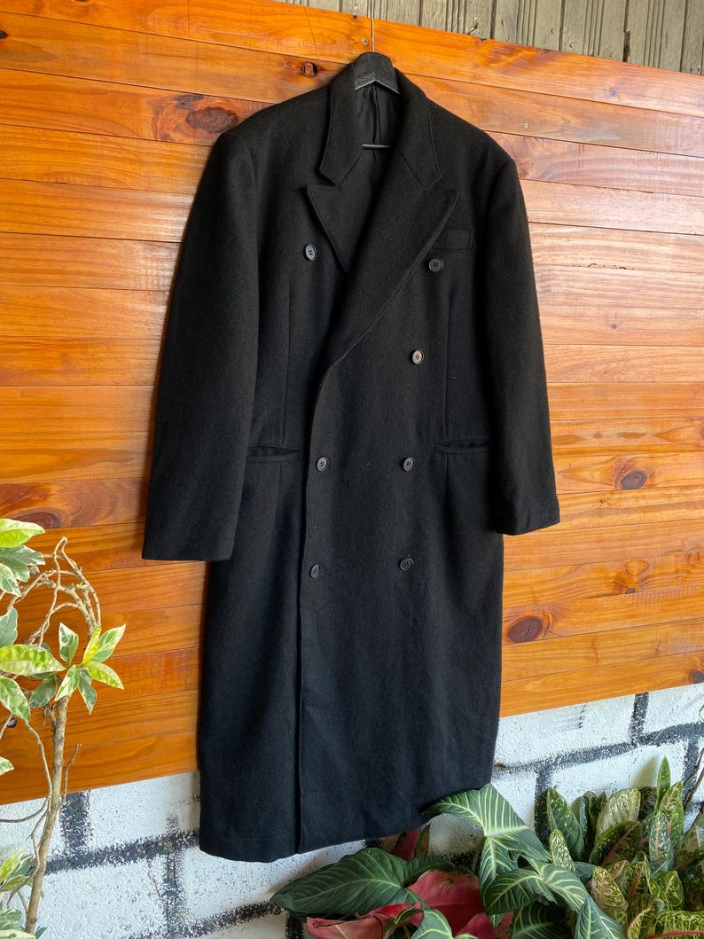 May include: A long, black double-breasted coat with a classic design. The coat features a notched lapel, multiple buttons, and a tailored fit. It is hanging on a black hanger against a wooden background.